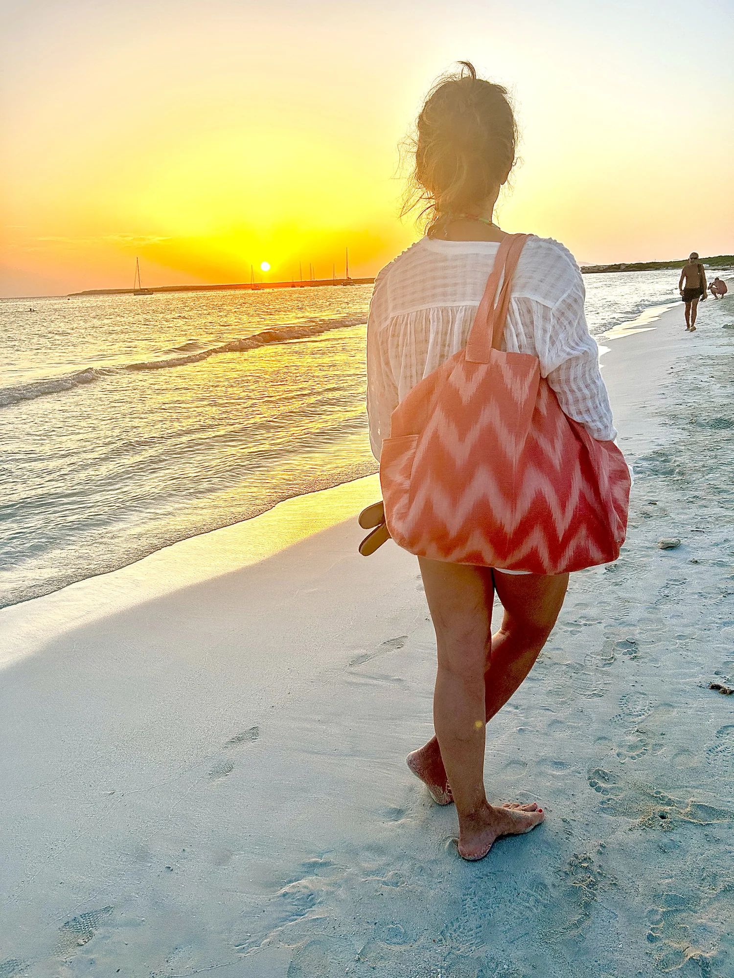 Mujer de espaldas caminando junto al mar al atardecer con una bolsa de tela de lenguas en tonos coral